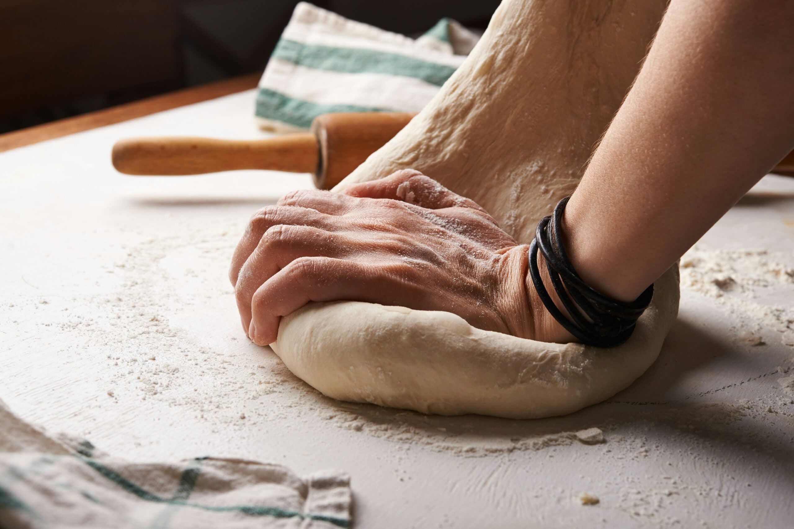 A close-up of a person’s hand kneading dough on a floured surface, with a rolling pin and striped kitchen towels nearby. The person is wearing dark bracelets on their wrist.