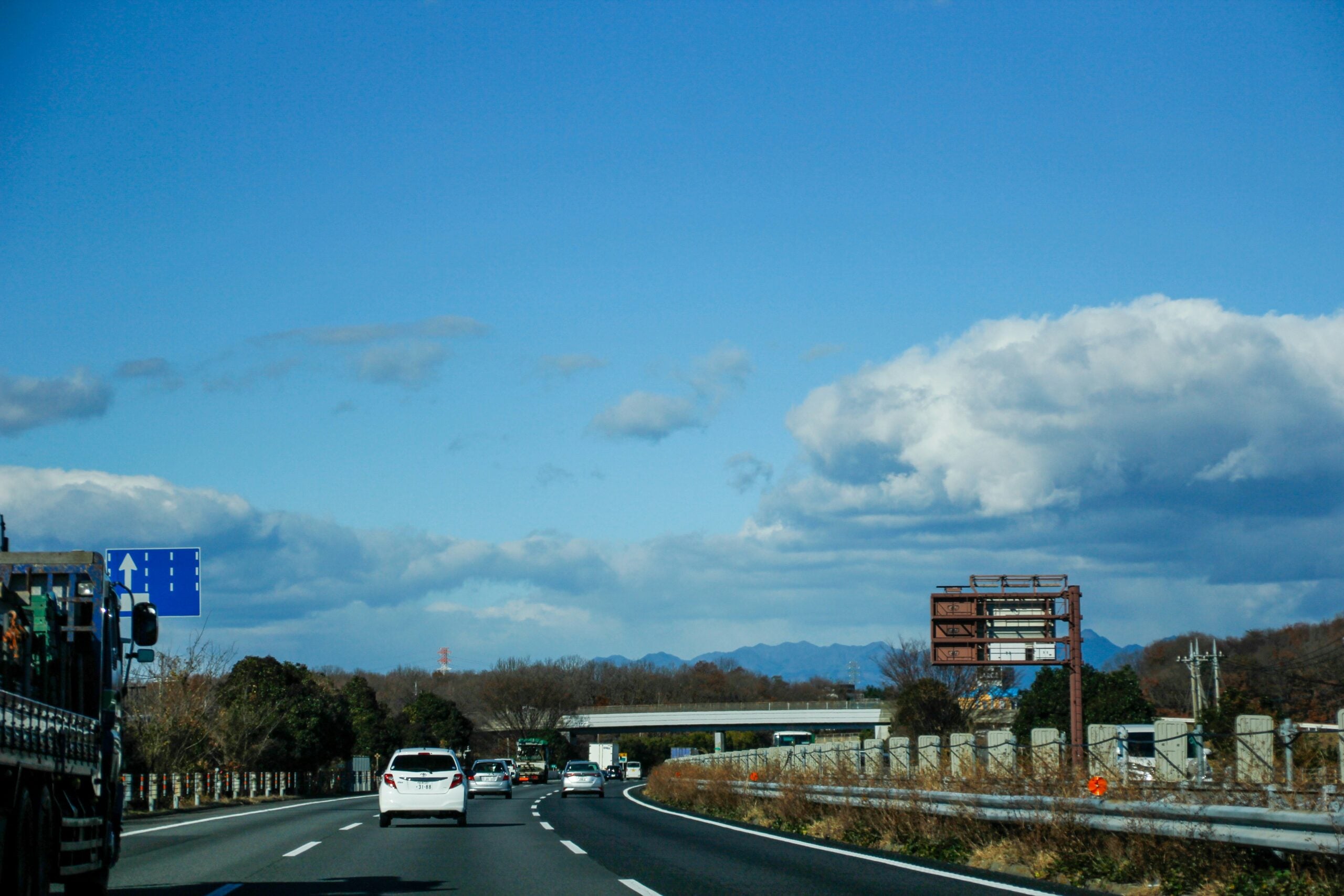 Cars driving on a multi-lane highway under a blue sky with scattered clouds, surrounded by trees and distant mountains. Road signs and a truck are visible on the left side of the image.
