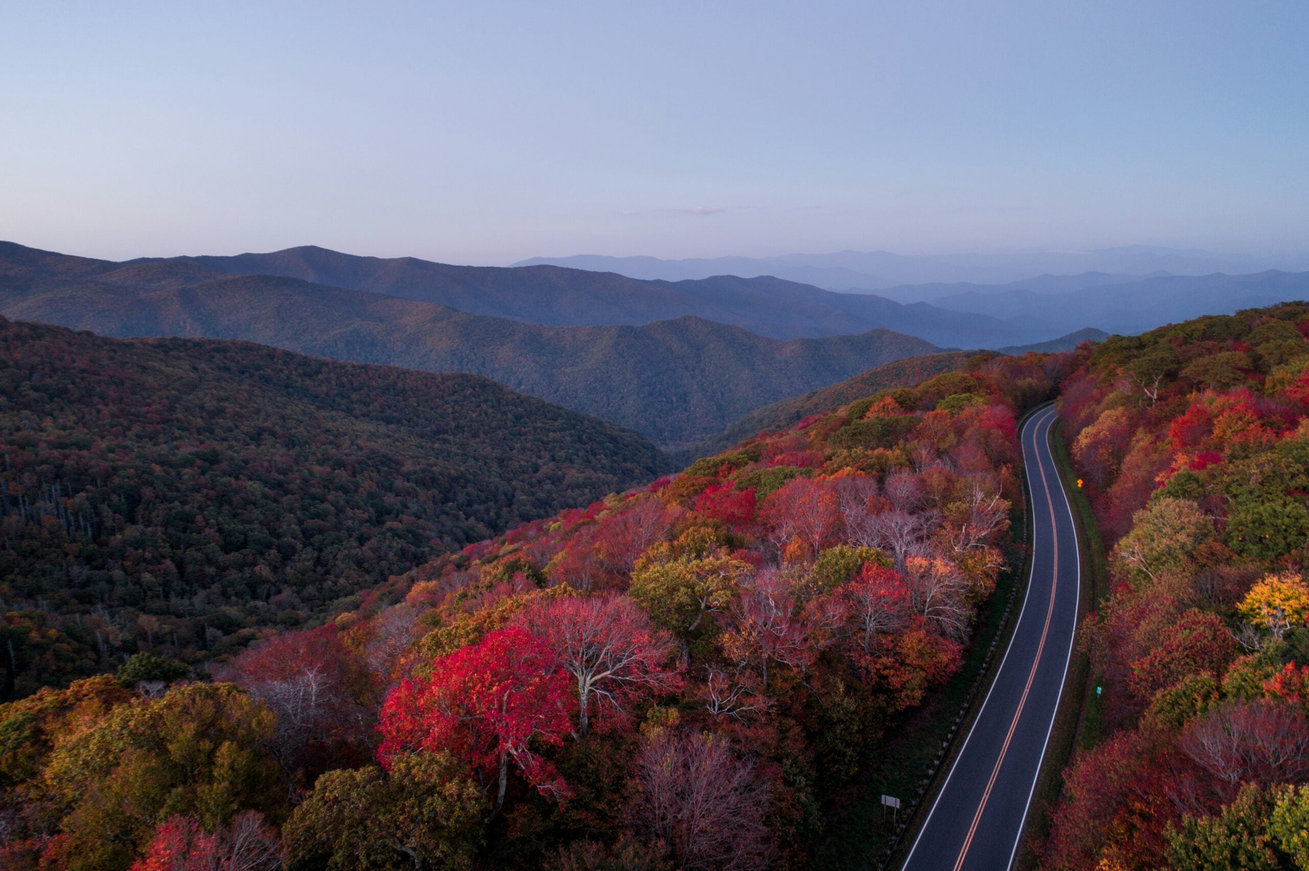 A winding road curves through hills covered in autumn foliage with vibrant red, orange, and green trees. Layers of distant mountains fade into the hazy blue sky at sunset.