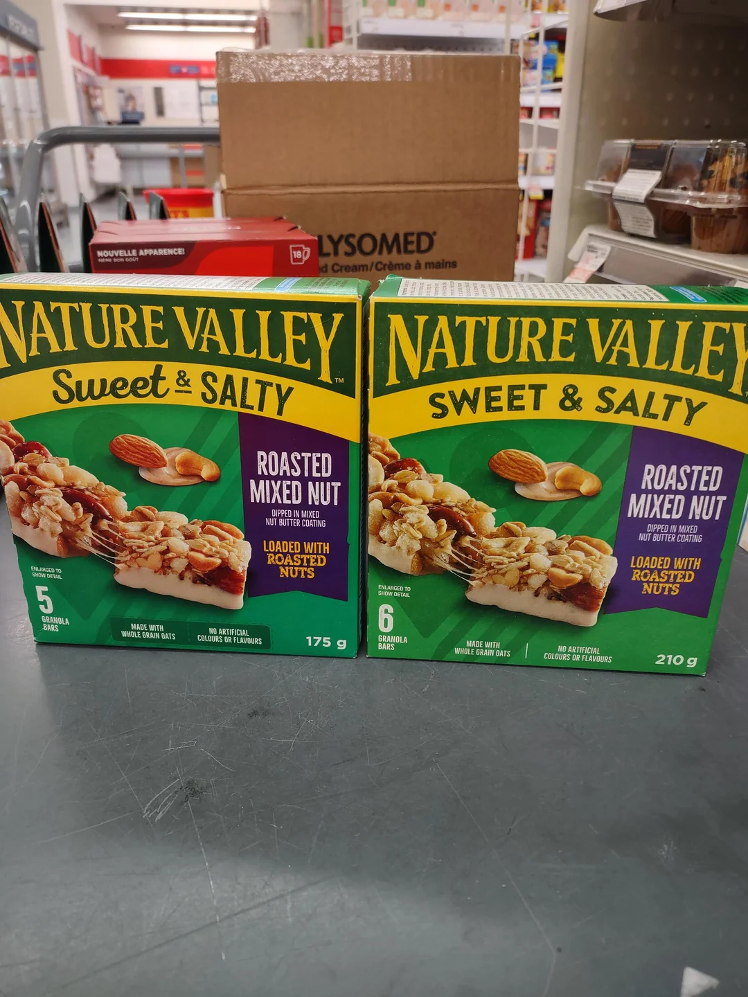 Two Nature Valley Sweet & Salty Roasted Mixed Nut granola bar boxes are side by side on a store counter. One box contains 5 bars (175g) and the other contains 6 bars (210g). Store shelves and products are in the background.