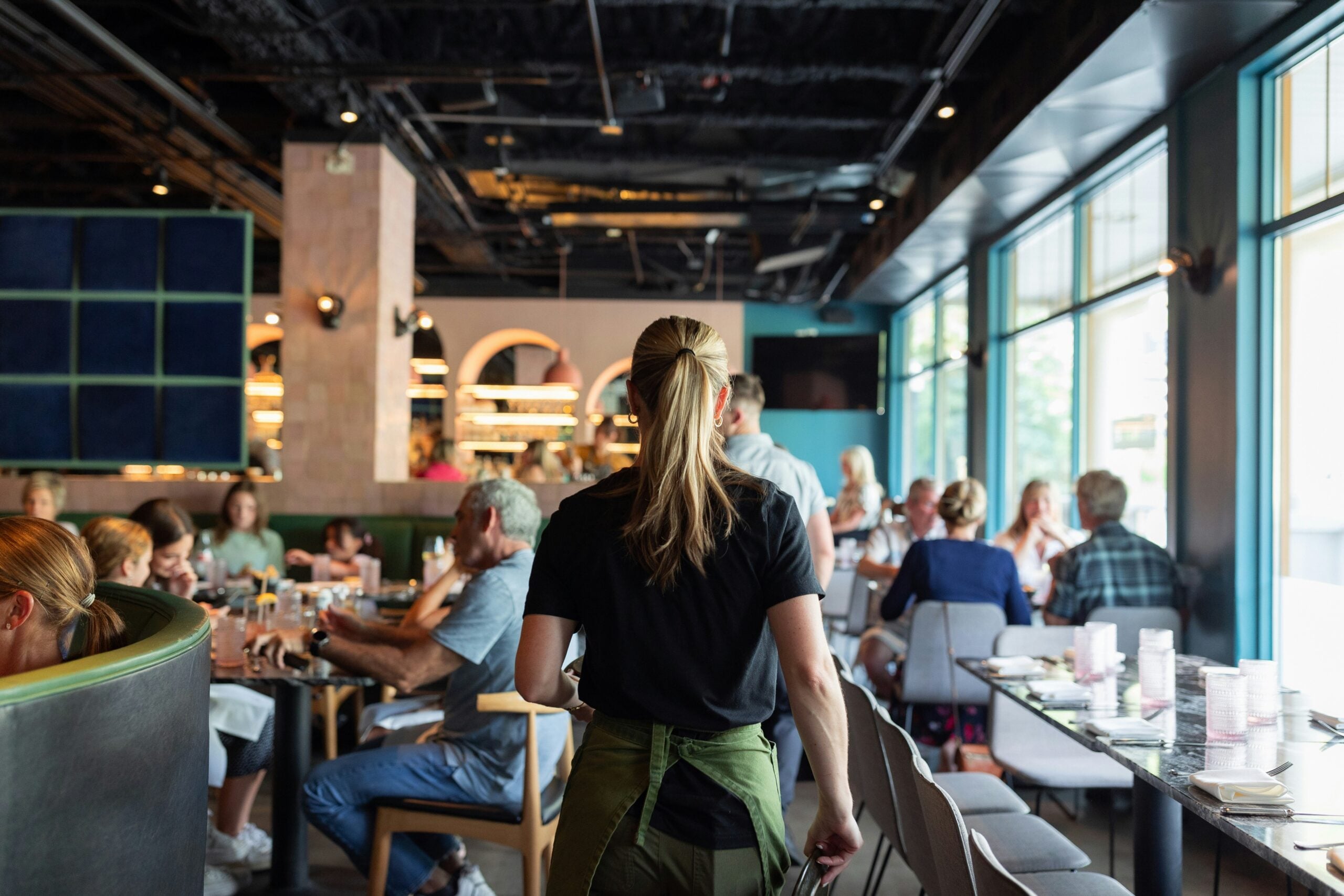 A server with a ponytail walks through a busy, modern restaurant filled with diners sitting at tables, chatting and eating. Large windows let in natural light, and the atmosphere is lively and casual.
