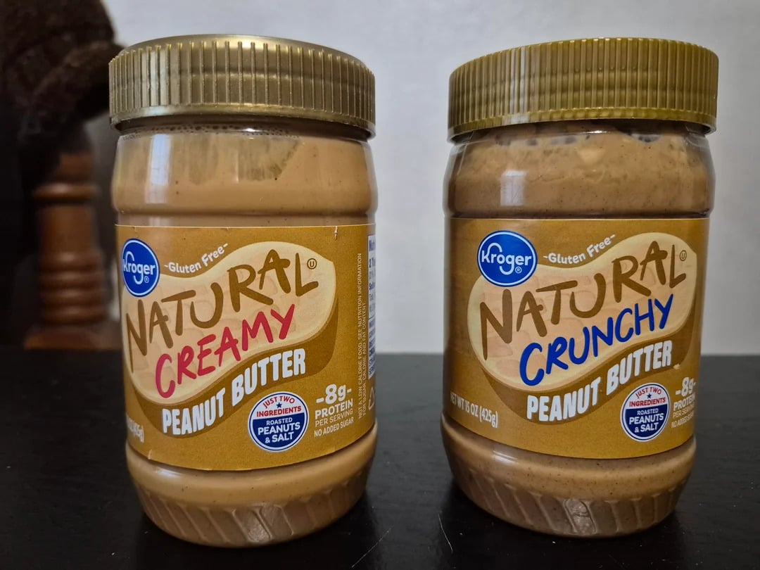 Two jars of Kroger Natural peanut butter, one Creamy and one Crunchy, are shown side by side on a dark surface—both feature gold lids and brown labels. Kroger is among the best peanut butter brands for rich flavor and variety.