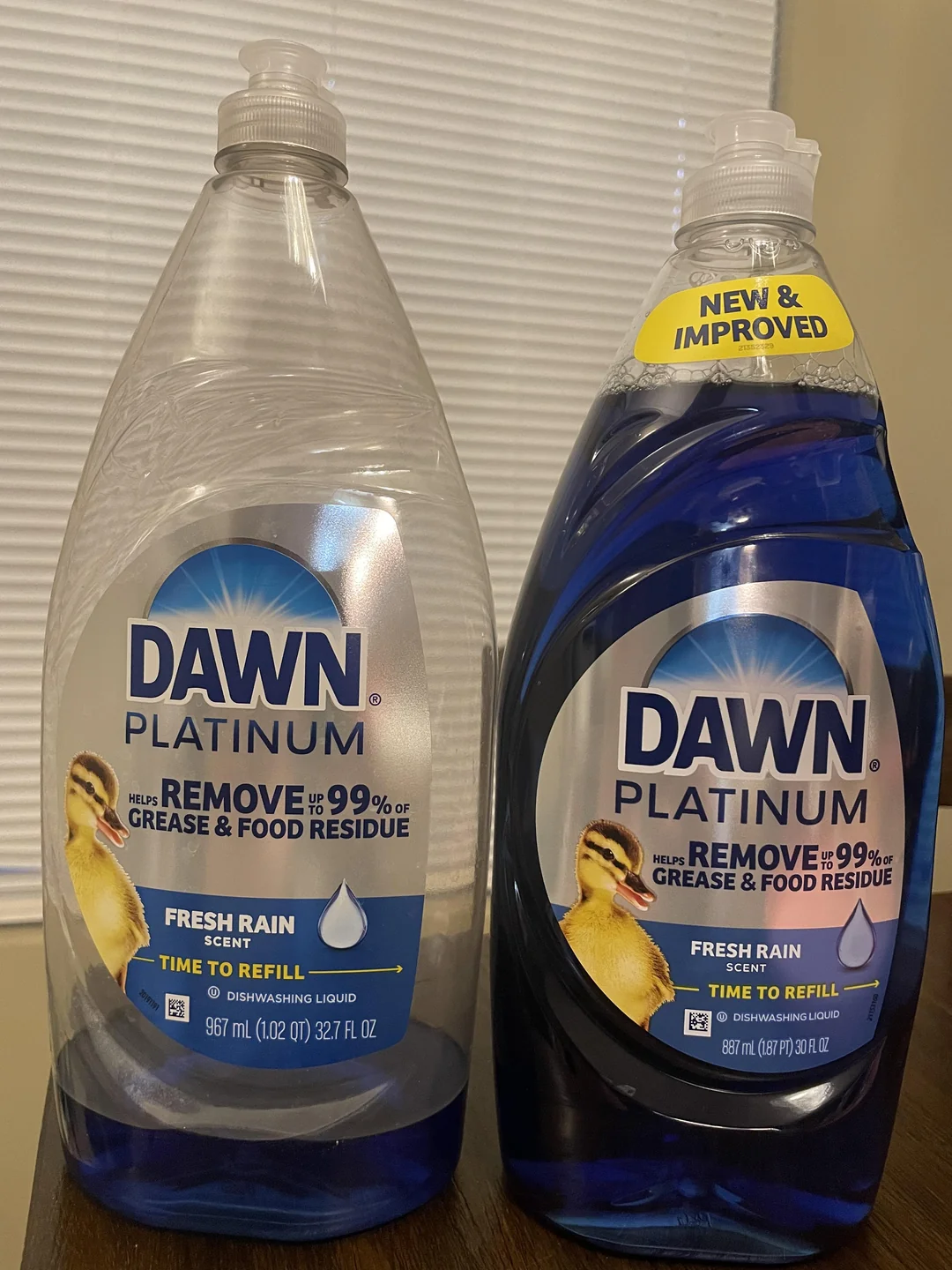 Two bottles of Dawn Platinum dishwashing liquid, both Fresh Rain scent. The left bottle is empty and clear; the right bottle is full with blue liquid and has a yellow "New & Improved" label on the cap.