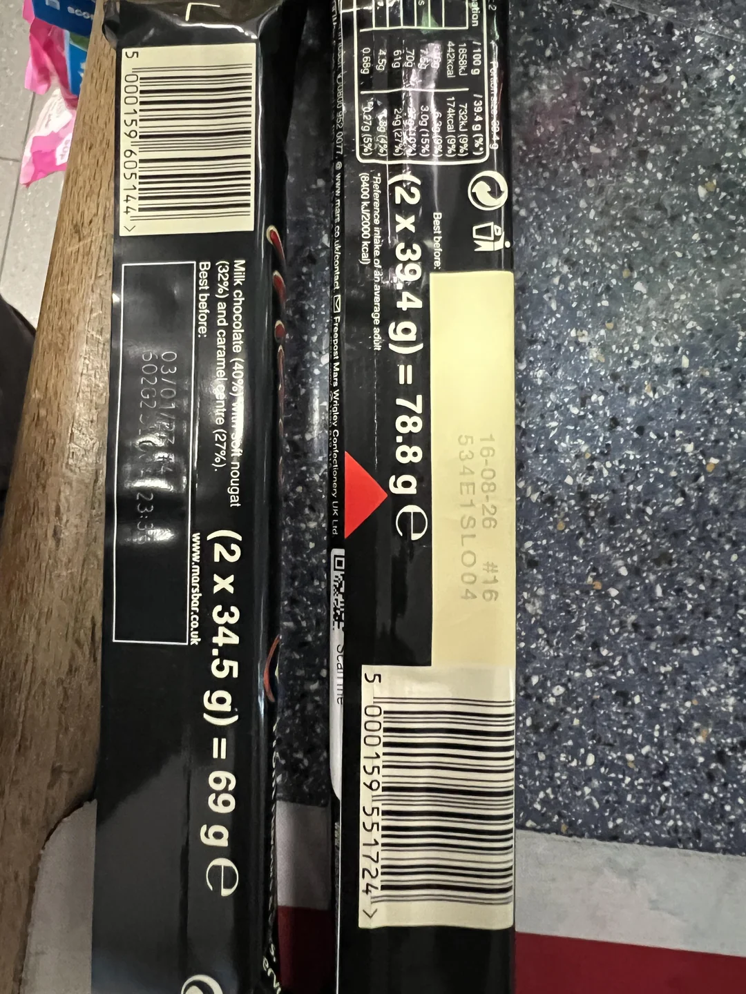 Two black packaged food items with barcodes and weight labels are placed on a dark countertop. One label reads "2x34.5g = 69g" and the other "2x39.4g = 78.8g." White and beige stickers with text are visible.