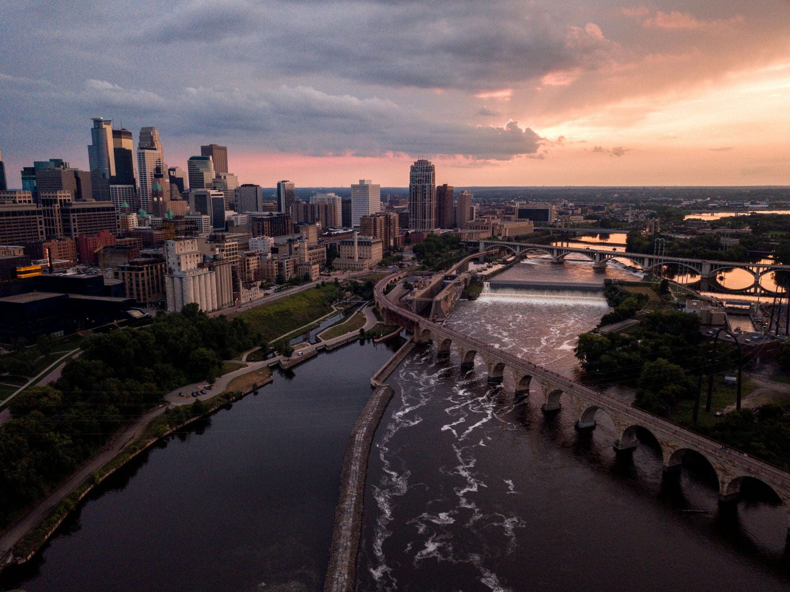 Aerial view of downtown Minneapolis at sunset, featuring skyscrapers, the Stone Arch Bridge crossing the Mississippi River, and a dramatic sky with clouds and soft sunlight.