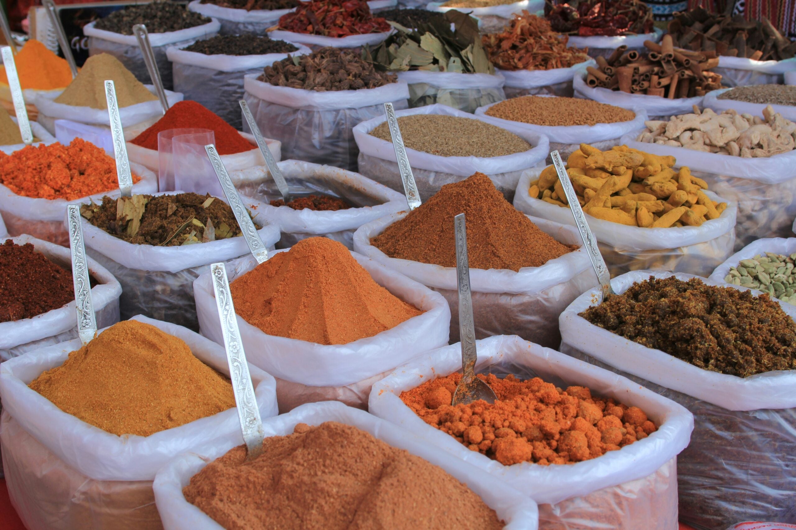 Bags filled with various colorful spices and herbs are displayed at an outdoor market, each with a metal scoop or spoon sticking out from the piles.