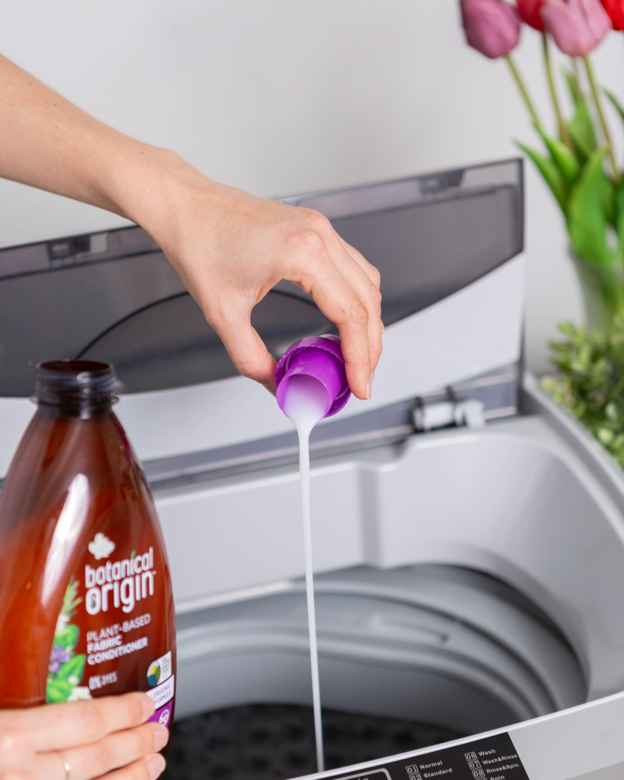 A person pours liquid laundry detergent from a bottle labeled "botanical origin" into a cap over an open top-loading washing machine. Pink tulips are visible in the background.
