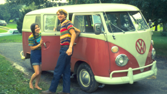 A smiling woman and man in striped shirts stand beside a vintage red and white Volkswagen van, capturing the charm of nostalgic photos on a driveway framed by green grass and trees.