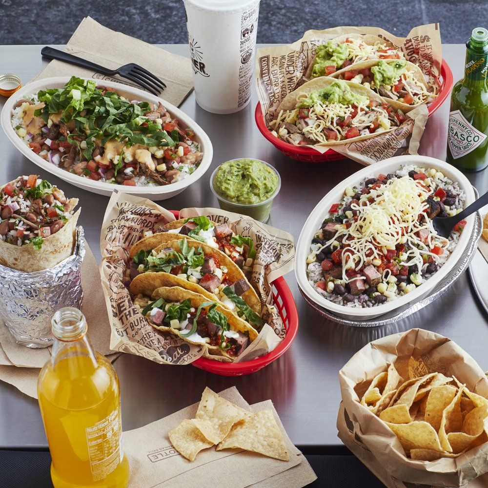 A table topped with various Mexican dishes including tacos, a burrito, a bowl with rice and beans, nachos with guacamole, and drinks like soda, a bottle of Jarritos, and a bottle of Tabasco sauce.