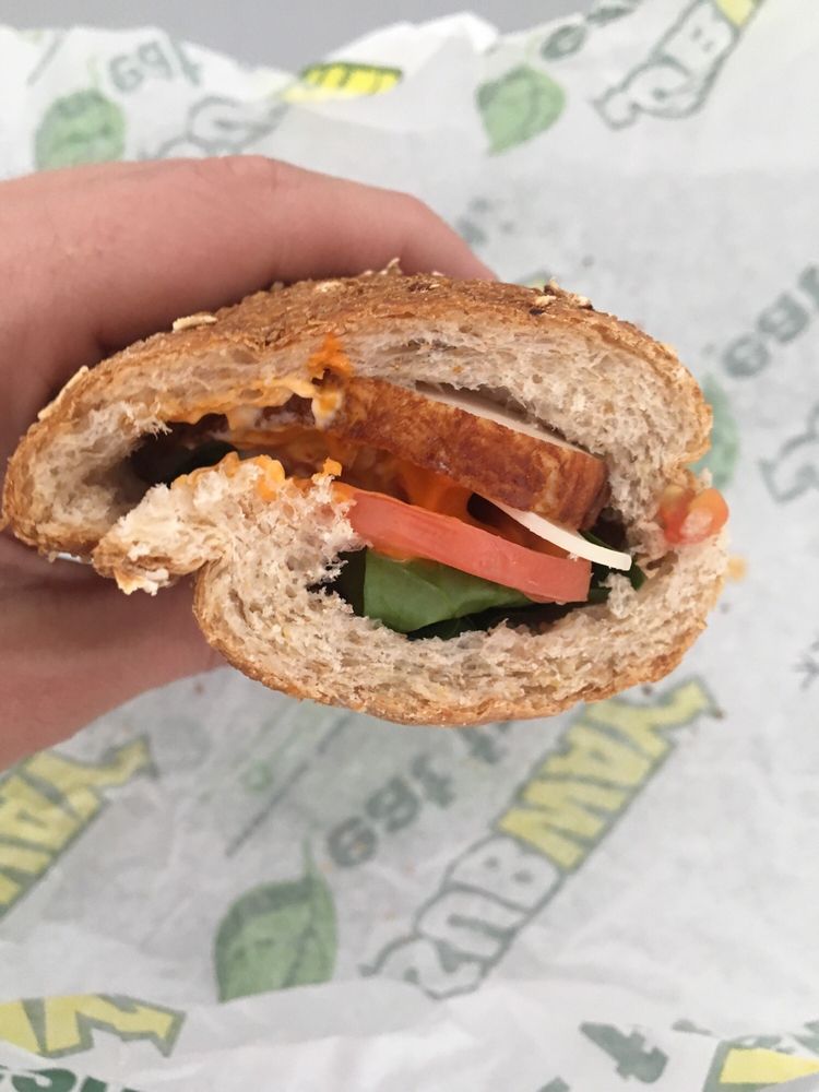 A hand holding a partially eaten sandwich on whole grain bread with visible slices of tomato, cucumber, cheese, and a brown filling. The sandwich rests on a Subway-branded sandwich wrapper.
