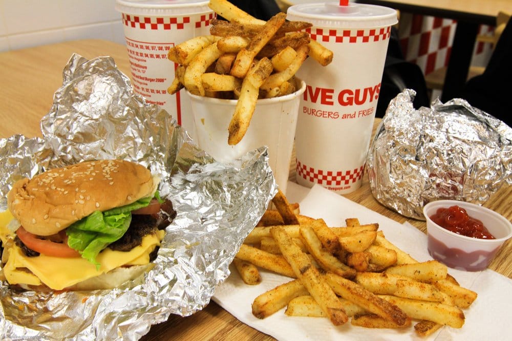 A cheeseburger with lettuce and tomato on foil, seasoned fries, cups of soda, and ketchup, all on a wooden table with "Five Guys" branding visible on the cups.
