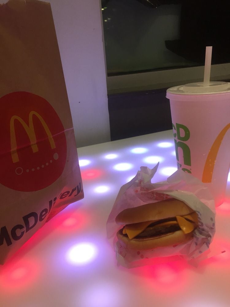 A McDonald's paper bag, a wrapped cheeseburger, and a large drink with a straw are placed on a table with glowing red and white lights.