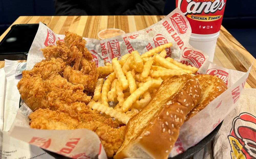 A tray of crispy chicken tenders, crinkle-cut fries, a slice of sesame seed toast, dipping sauce, and a soft drink on a wooden table, with Raising Cane’s branded paper and cup.