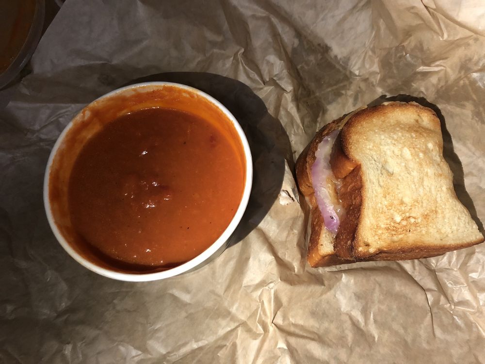 A bowl of tomato soup sits next to a grilled cheese sandwich on crumpled brown paper.