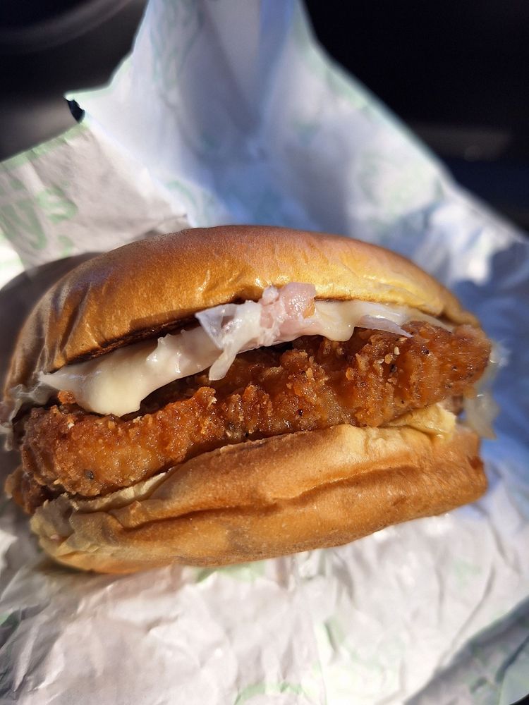 A close-up of a crispy Chicken Sandwich with mayonnaise and shredded lettuce on a soft, toasted bun, partially wrapped in white paper.