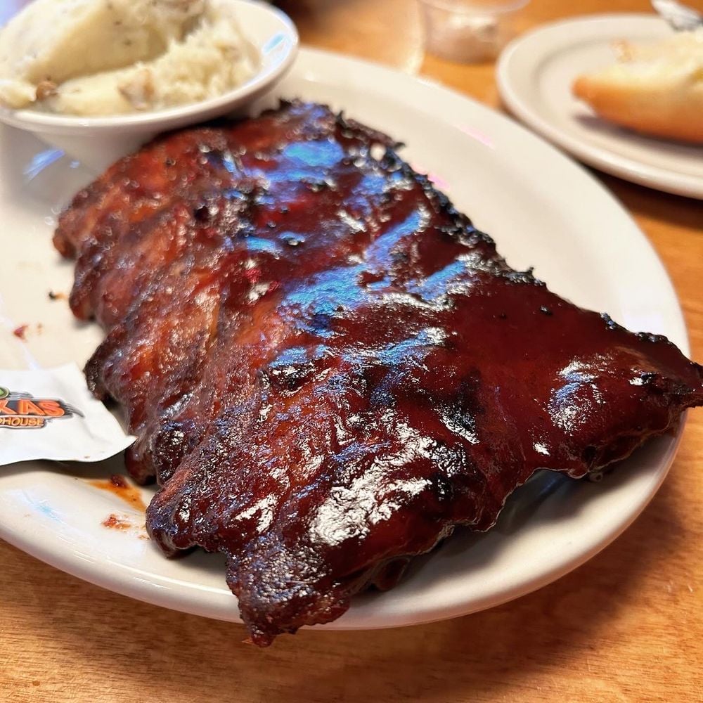 A plate of glazed barbecue ribs is served with a bowl of mashed potatoes on the side. The food is on a white plate atop a wooden table, with a piece of bread visible in the background.