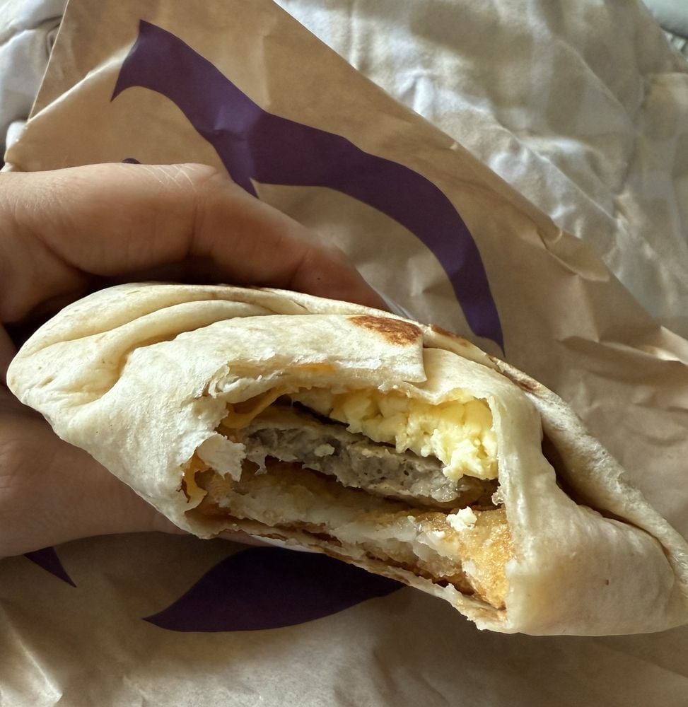 A hand holds a partially unwrapped breakfast wrap showing layers of scrambled eggs, sausage patty, and hash brown inside a soft tortilla. The wrap is on crumpled paper packaging with a purple design.