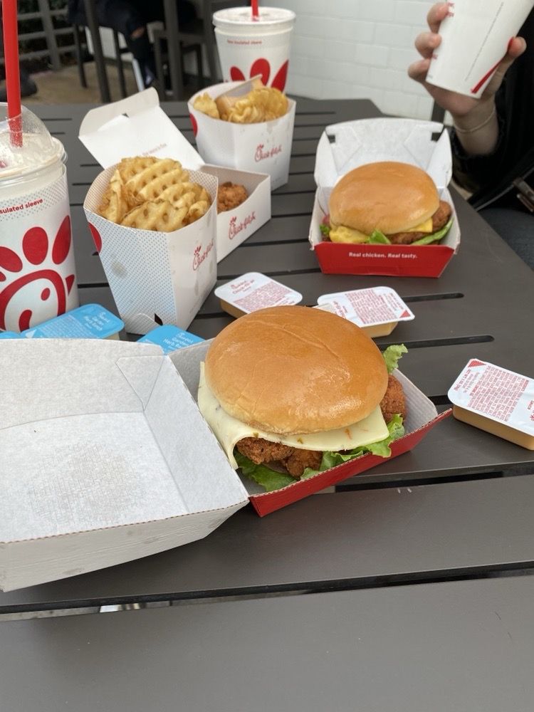 Three Chick-fil-A chicken sandwiches, waffle fries, chicken nuggets, dipping sauces, and two drinks with red straws are on a gray outdoor table. A person is holding one of the drinks in the background.