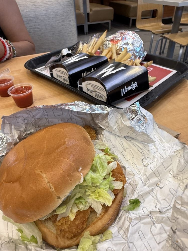 A close-up of a chicken sandwich with lettuce on a wrapper sits on a table, with a tray in the background holding two wrapped burgers, fries, and small cups of ketchup at a Wendy’s restaurant.