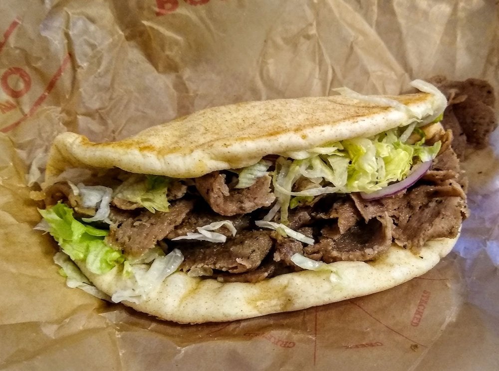 A gyro sandwich with sliced seasoned meat, shredded lettuce, and red onions, served in a folded pita bread on brown deli paper.