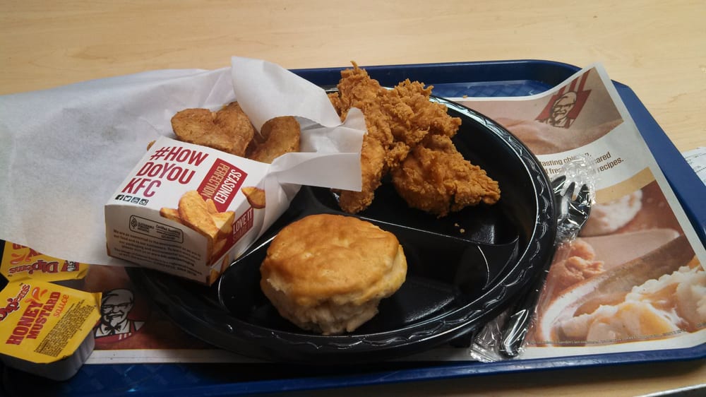 A KFC meal on a blue tray featuring fried chicken, potato wedges, a biscuit, a container of honey sauce, packaged utensils, and KFC-branded paper items in the background.