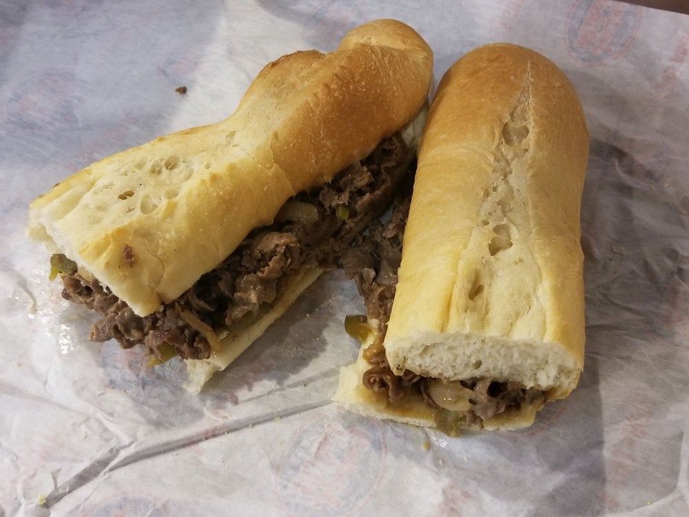 A sandwich with sliced steak, onions, and peppers on a hoagie roll, cut in half and placed on deli paper.
