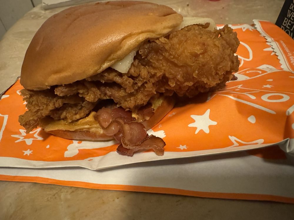 A close-up of a fried chicken sandwich with bacon and cheese on a bun, sitting on an orange and white wrapper on a countertop.