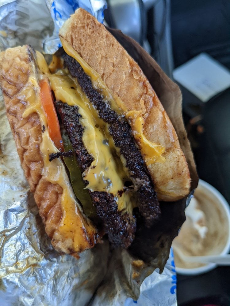 A close-up of a cheeseburger with two beef patties, melted cheese, pickles, tomato, and a toasted bun, partially wrapped in foil. A cup with a drink or dessert is visible in the background.