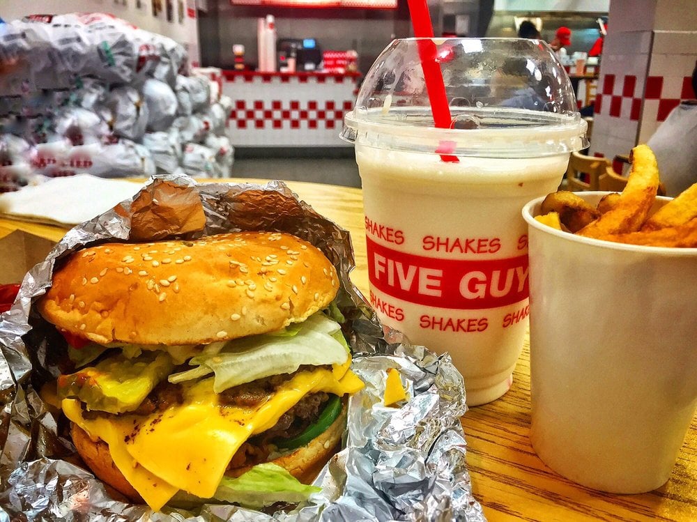 A cheeseburger in foil, a cup of fries, and a milkshake with a red straw on a wooden table inside a Five Guys restaurant; potato sacks are visible in the background.