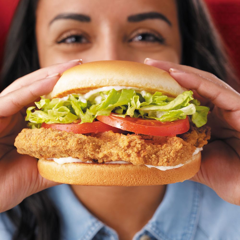 A person holding a delicious Chicken Sandwich with lettuce, tomato, and mayonnaise on a bun, smiling in the background.