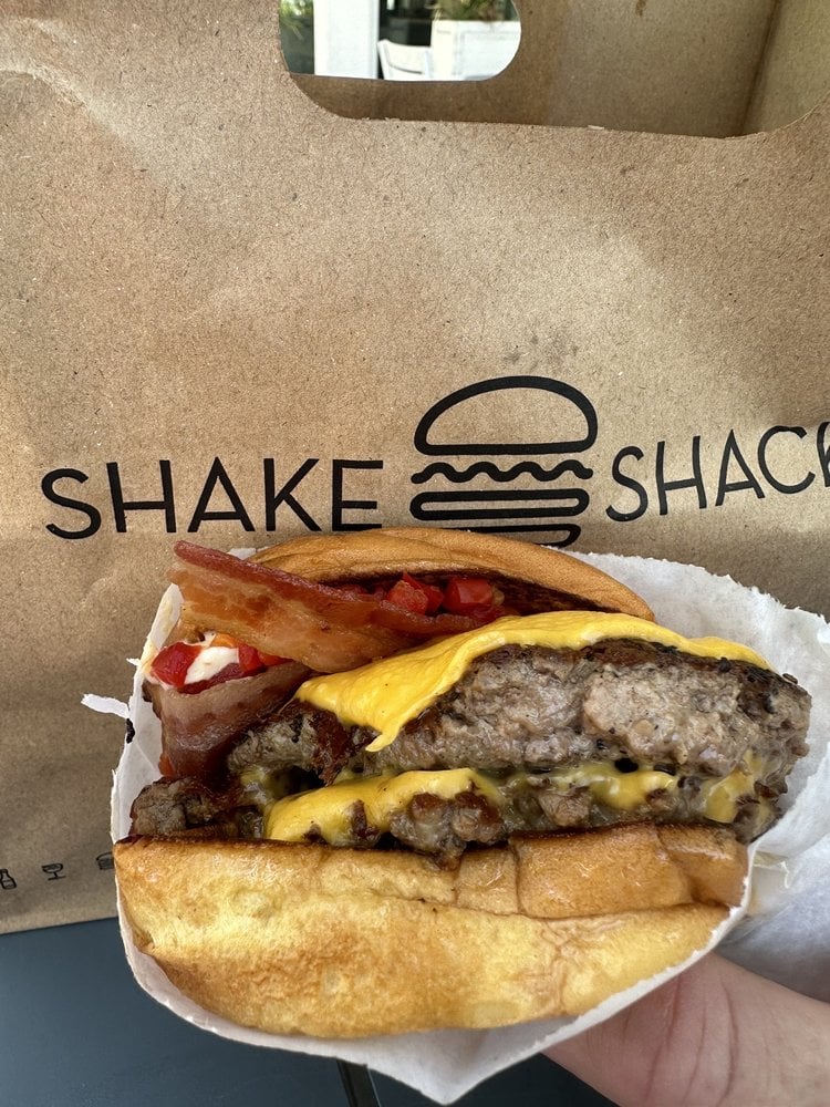 A hand holds a cheeseburger with bacon and red peppers in front of a brown Shake Shack paper bag with a burger logo and the words "SHAKE SHACK" printed on it.