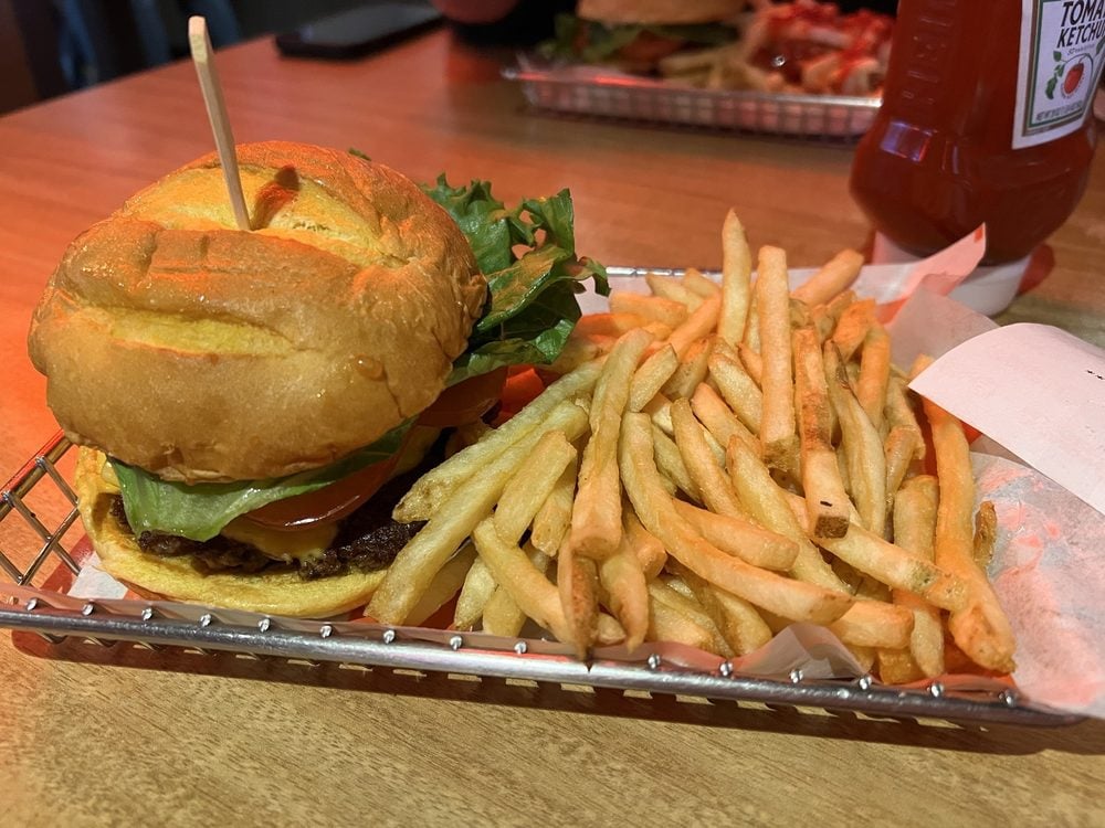 A tray with a cheeseburger topped with lettuce and tomato, and a generous serving of French fries on the side. Ketchup bottle and another tray of food are visible in the background on a wooden table.