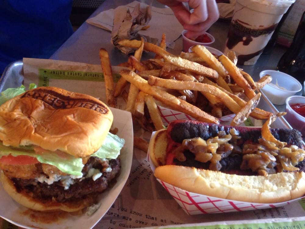A tray with a cheeseburger topped with lettuce and tomato, a grilled hot dog with onions, and a generous serving of French fries. A hand reaches for fries, and milkshakes and sauces are visible in the background.