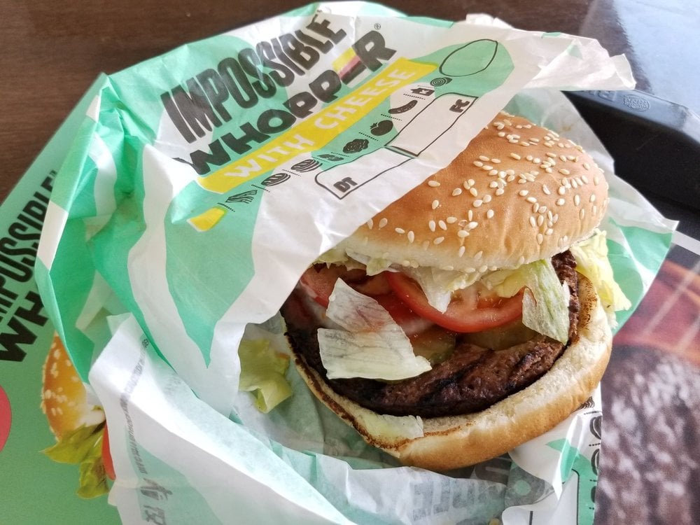 An Impossible Whopper with cheese from Burger King, partially unwrapped, showing a sesame seed bun, lettuce, tomato, pickles, cheese, and a plant-based patty on a wrapper.