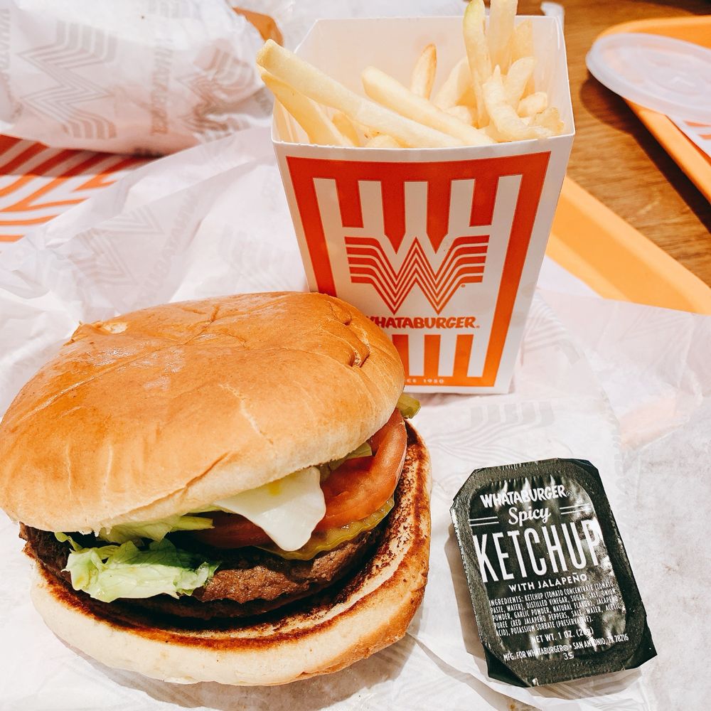 A Whataburger meal with a hamburger, a container of French fries in orange-striped packaging, and a packet of spicy ketchup with jalapeño on white paper wrappers.