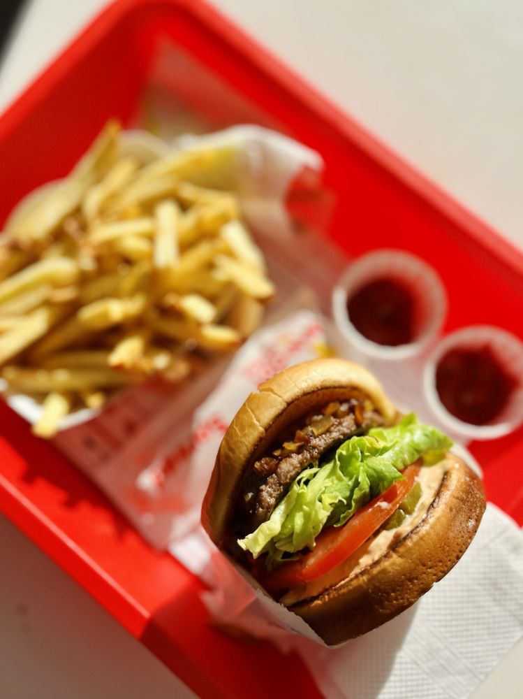 A red tray holds a burger with lettuce, tomato, and sauce, alongside a serving of French fries and two small cups of ketchup.
