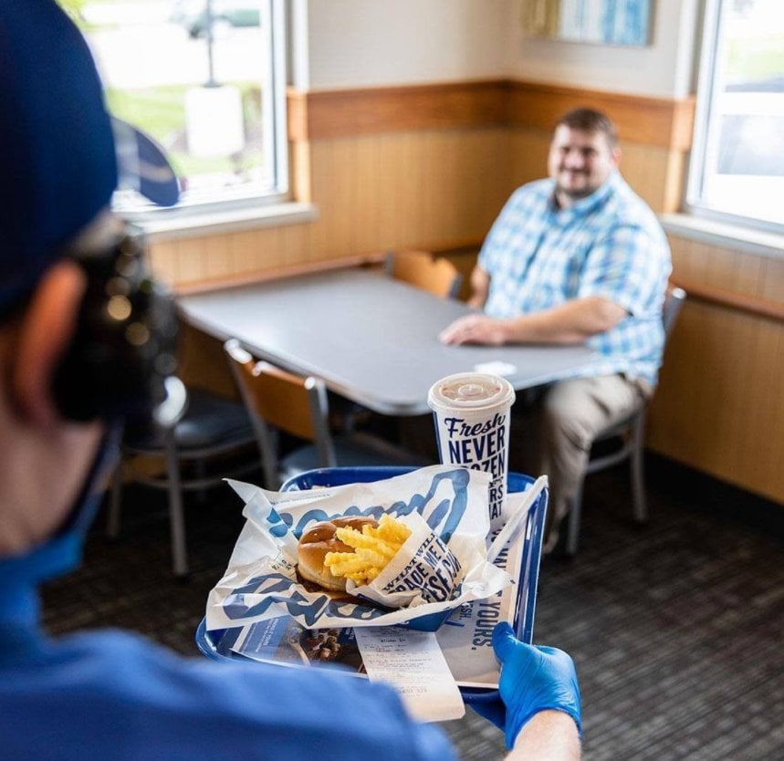 A server wearing blue gloves carries a tray with fries, a burger, and a drink toward a smiling man sitting alone at a table in a bright, casual restaurant.