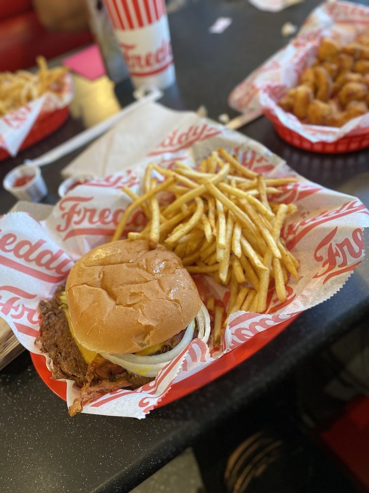 A tray with a cheeseburger topped with onions and pickles, a large serving of thin-cut French fries, all on red and white Freddy’s branded paper. Other food trays and a drink cup are visible in the background.