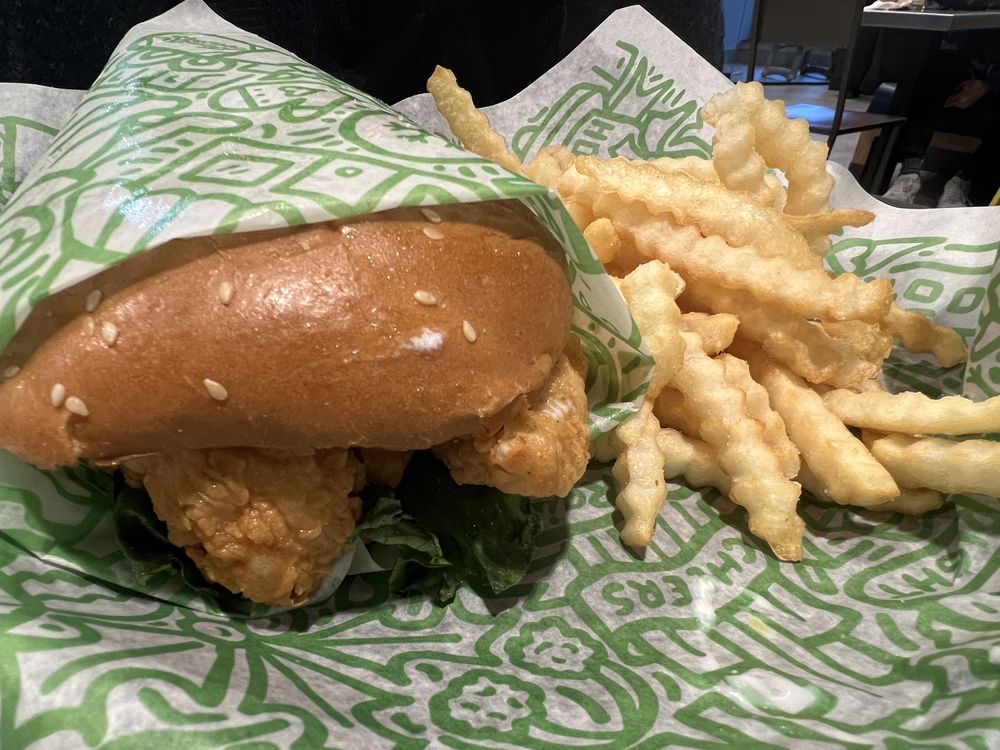 A close-up of a Chicken Sandwich with crispy fried chicken and lettuce in a sesame seed bun, served alongside crinkle-cut French fries on green and white patterned paper.