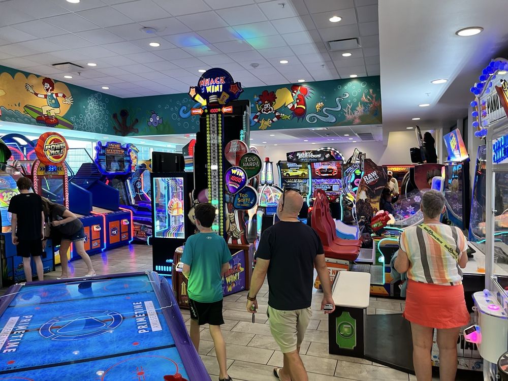 People walk and play various colorful arcade games in a lively indoor arcade with bright lighting, cartoon murals on the walls, and multiple game machines—reminiscent of some of the coolest McDonald's locations around the world.