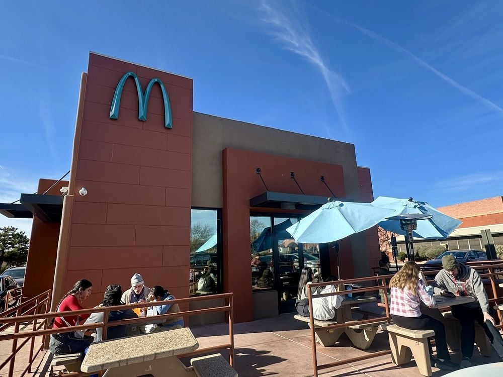 People sit at outdoor tables under blue umbrellas outside a McDonald's restaurant on a sunny day. With its modern design and iconic yellow "M" logo, this spot stands out as one of the coolest McDonald's locations to visit.