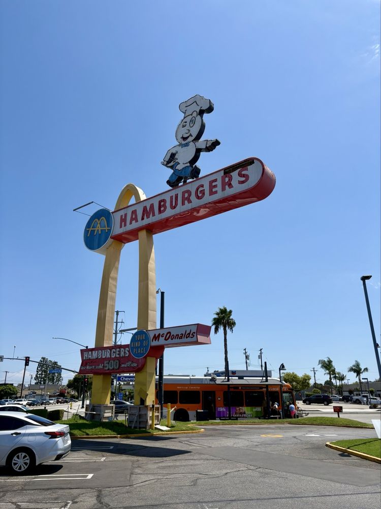 A vintage McDonald’s sign featuring a chef character holding a "HAMBURGERS" sign stands tall above a parking lot with cars and a McDonald’s restaurant building, making it one of the Coolest McDonald's Locations on a sunny day.