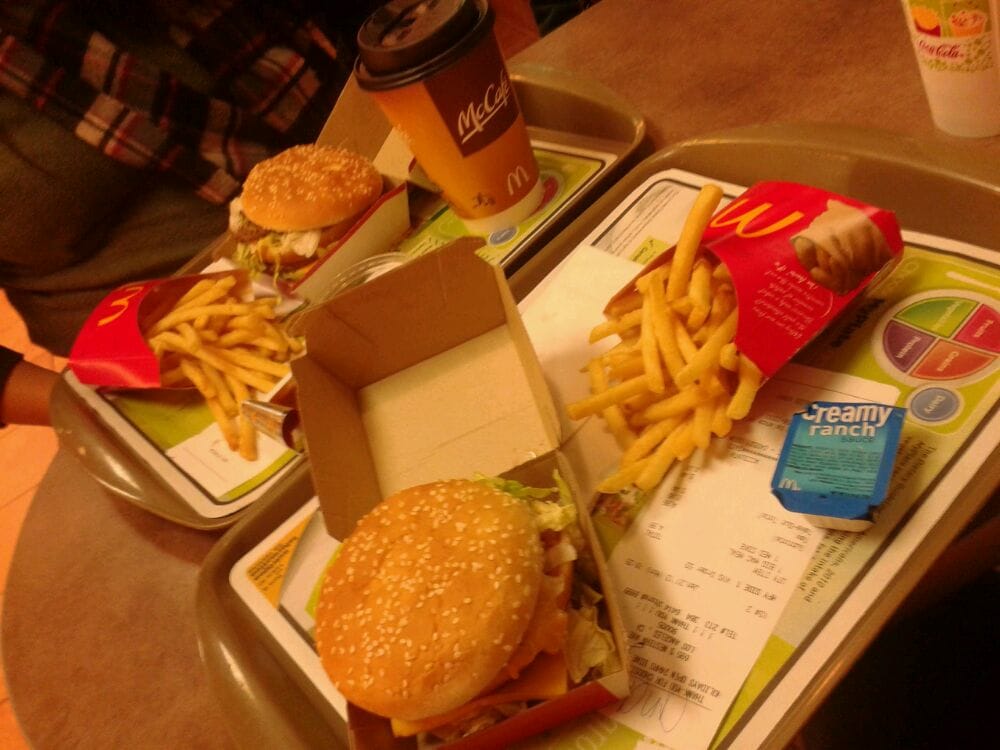 Two trays of McDonald's food on a table, each with a burger, fries, and a drink. One tray has a coffee cup, the other has a cold drink and a packet of creamy ranch sauce. A person in a plaid shirt sits nearby.