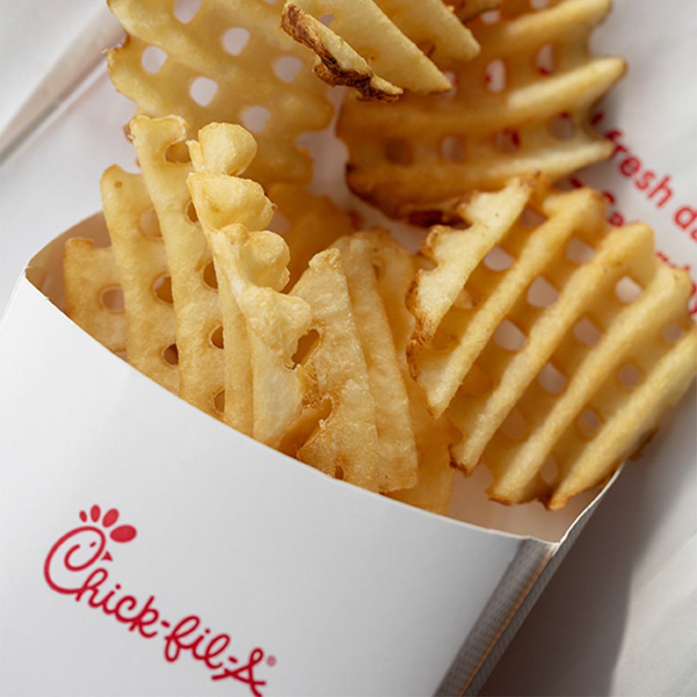 A close-up of waffle fries in a white Chick-fil-A container, showing the brand’s red logo and crispy, golden fries with a grid pattern.