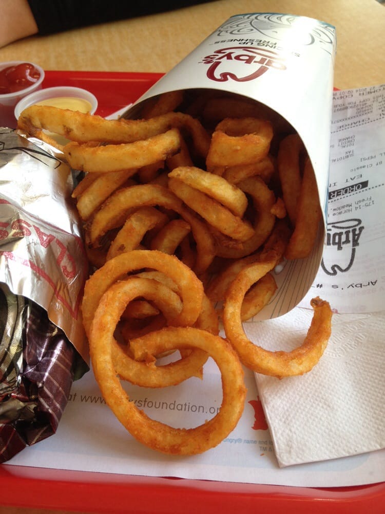 A serving of curly fries spilling out of Arby’s branded packaging onto a tray, next to a foil-wrapped sandwich, ketchup cup, napkin, and receipt.