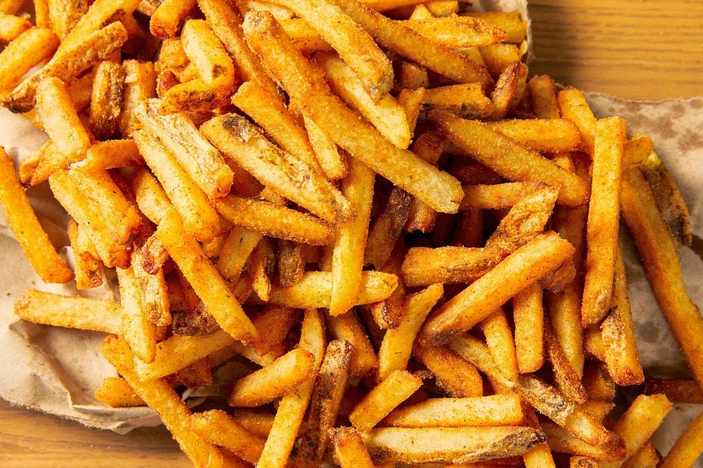 A close-up view of a large pile of seasoned French fries on brown parchment paper, showcasing their golden, crispy texture and dusting of spices.