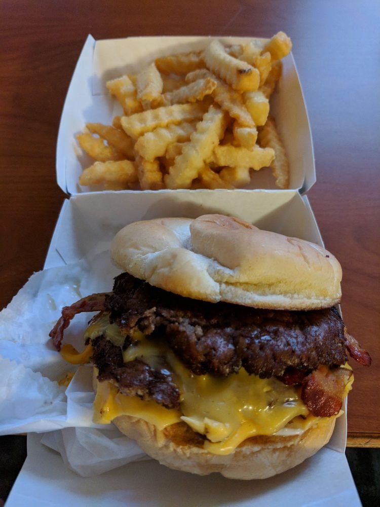 A cheeseburger with bacon, melted cheese, and a toasted bun sits in a takeout container next to a serving of crinkle-cut French fries on a wooden table.