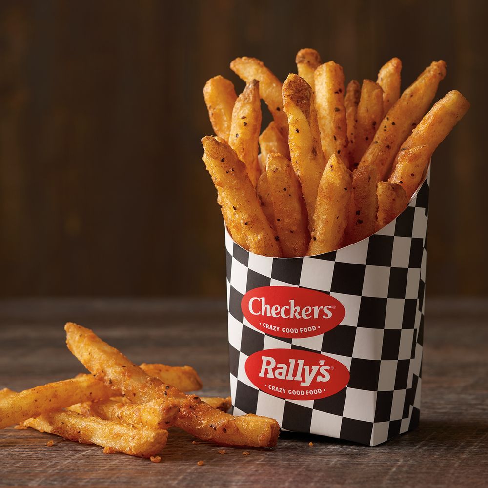 A black-and-white checkered container filled with seasoned French fries, labeled with "Checkers" and "Rally's" logos, sits on a wooden surface with a few fries scattered nearby.