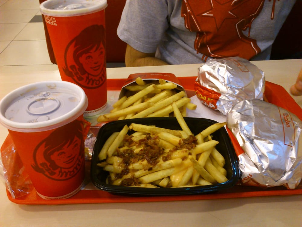 A Wendy’s meal on a tray includes two burgers wrapped in foil, two servings of fries (one with cheese and bacon), and two soft drinks in red Wendy’s cups. A person wearing a red shirt sits behind the tray.