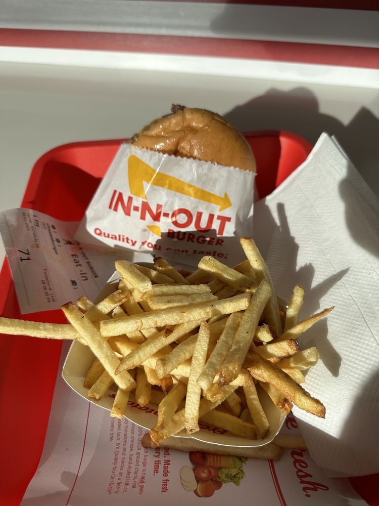 A tray with an In-N-Out burger in a wrapper and a container of thin, golden French fries sits on a table, partially covered by a napkin, with sunlight casting shadows across the food.